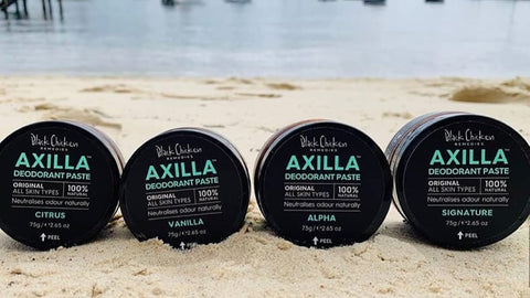 Range of Black Chicken Remedies Axilla Natural Deodorant Paste lying in a row face-forward on a beach. 