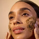 Woman applying a facial scrub to her face with a beige background