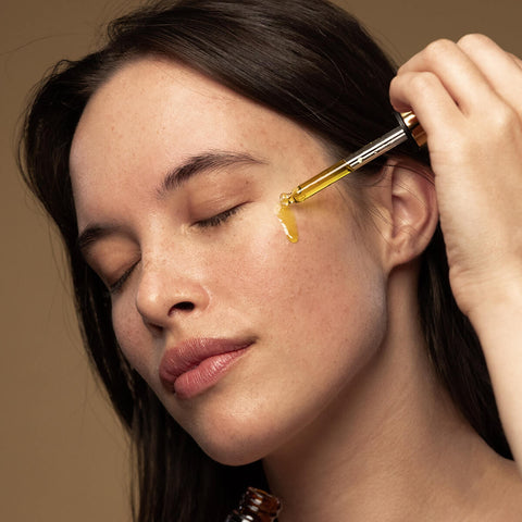 Model using applicator to drop some face oil on her cheek.