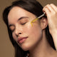 Model using applicator to drop some face oil on her cheek.