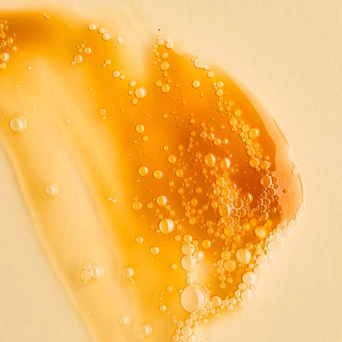 Swatch of Juliet Anti-Blemish Gel Cleanser with caramel colouring and light texture with bubbles on a pale yellow background.
