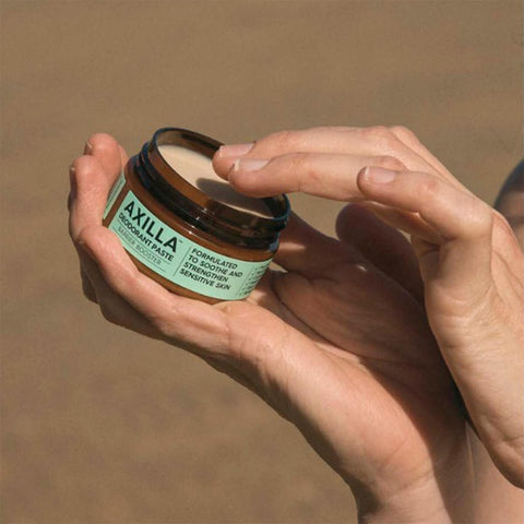 Black Chicken Remedies Axilla Deodorant Paste Barrier Booster lifestyle close up of hands opening the tin to show the paste, with a beach behind
