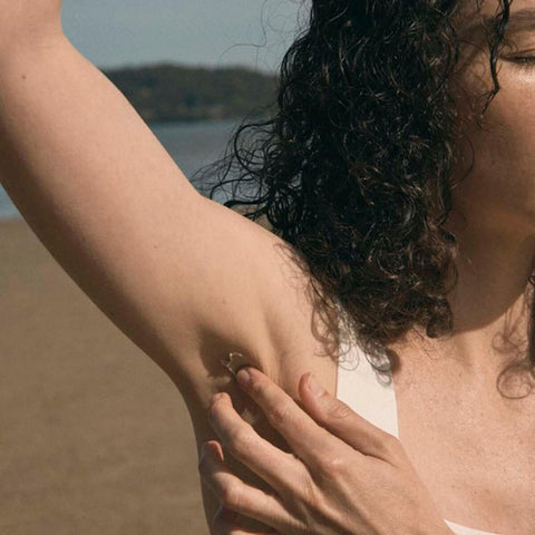 Black Chicken Remedies Axilla Deodorant Paste Original lifestyle shot of a woman with dark curly hair raising her arm to apply the paste on a beach