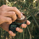 Black Chicken Remedies Axilla Deodorant Paste Original lifestyle close up of hands opening the tin outdoors with tall grass behind