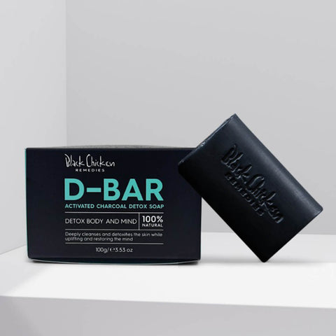 Black Chicken Remedies D-Bar Activated Charcoal Detox Soap 100g lifestyle shot of the black box and a charcoal soap bar on a pale shelf