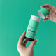 Black Chicken Remedies Dry Shampoo lifestyle shot of hands holding the green stick and lifting off its cap against a bright green backdrop