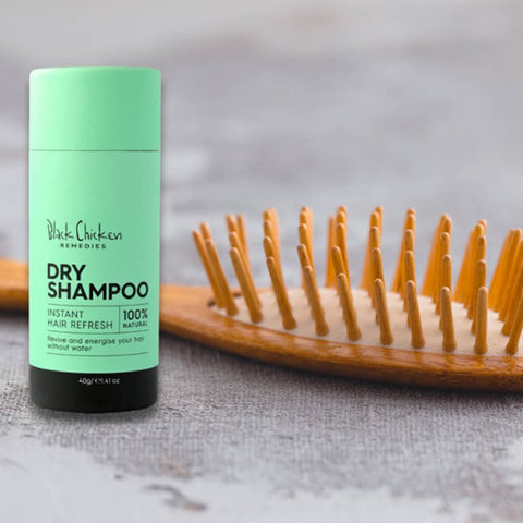 Black Chicken Remedies Dry Shampoo lifestyle shot of the green stick standing beside a wooden hairbrush on a grey surface, close up