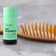 Black Chicken Remedies Dry Shampoo lifestyle shot of the green stick standing beside a wooden hairbrush on a grey surface, close up