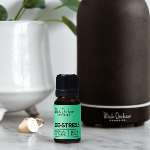 Black Chicken Remedies Essential Oil Blend De-Stress 9ml lifestyle shot of the small bottle in front of a dark diffuser and a white vase