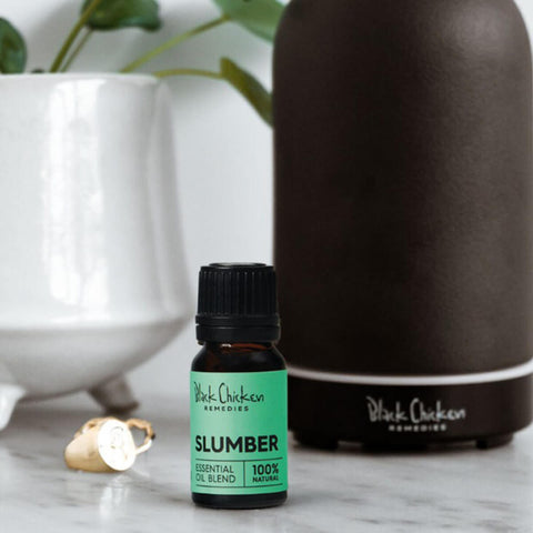 Black Chicken Remedies Essential Oil Blend Slumber lifestyle shot of the small bottle in front of a dark diffuser and a white vase