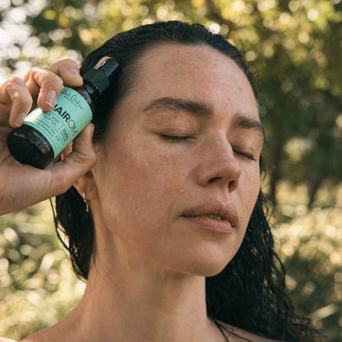 Black Chicken Remedies HairOM Hair & Scalp Oil 50ml lifestyle shot of a woman with wet dark hair applying the oil to her scalp, trees behind her