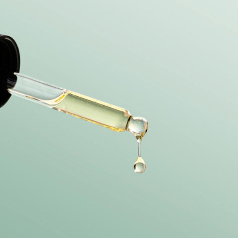 Black Chicken Remedies Love Your Face Serum lifestyle shot of a glass dropper over a pale green backdrop with a drop of serum falling