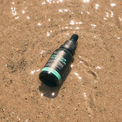 Black Chicken Remedies Love Your Face Serum lifestyle shot of the dropper bottle lying in shallow water over sand, with sunlight on the ripples