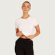 A woman modeling a fitted white short-sleeve crop top and black leggings against a light gray studio background. The top is slightly cropped at the hem, and she is lifting the fabric with both hands.