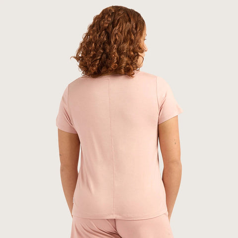 A woman with curly brown hair is shown from the back wearing a light pink short-sleeve top and matching pink bottoms against a light gray studio background.