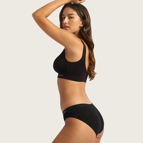 Boody Hipster Bikini Black lifestyle shot of a woman with long dark hair turned side on, one arm raised, in the black crop top and briefs