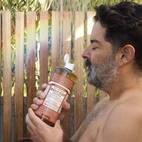 Man holding a bottle of Dr. Bronner's 18-in-1 Pure-Castile Magic Soap -Eucalyptus outdoors with a wooden fence in the background.