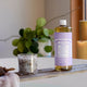 Bottle of Dr. Bronner's 18-in-1 Pure-Castile Magic Soap - Lavendar on a surface with a candle & plant in the background.