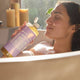 Woman in a bathtub holding a bottle of 
Dr. Bronner's 18-in-1 Pure-Castile Magic Soap - Lavender with candles in the background.