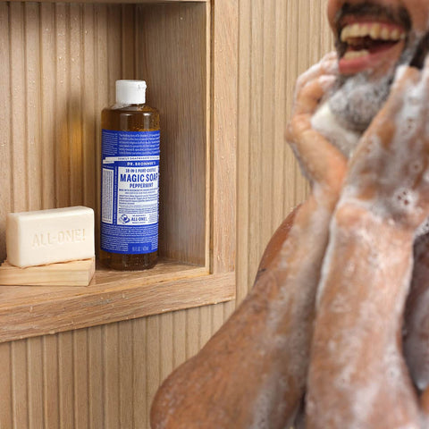 Person washing their face with lathered soap bubbles with a bottle of Dr. Bronner's Magic Soap on a wooden shelf.
