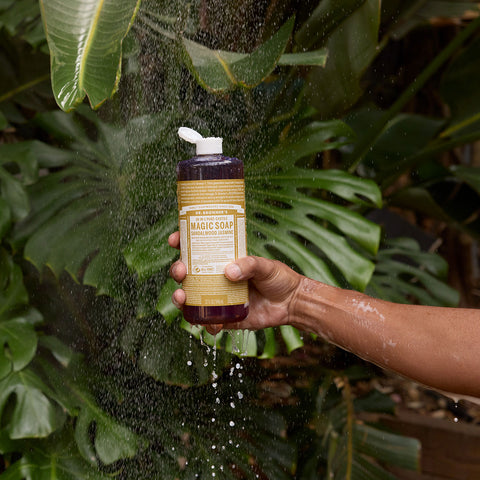 Person holding a bottle of Dr. Bronner's 18-in-1 Pure-Castile Magic Soap - Sandalwood Jasmine under a shower with green leaves in the background.