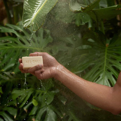 Hand holding a bar of soap under a shower with green leaves in the background