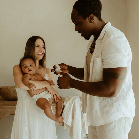 Woman holding a baby while a man holding a baby wipe stands nearby in a softly lit room.