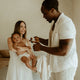 Woman holding a baby while a man holding a baby wipe stands nearby in a softly lit room.