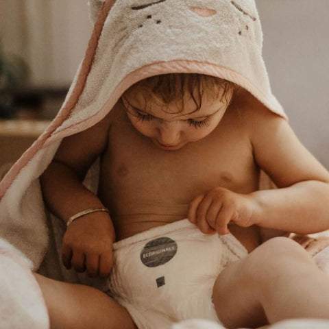 Baby wrapped in a towel, wearing a pull-up with a brand label, sitting on a soft surface.