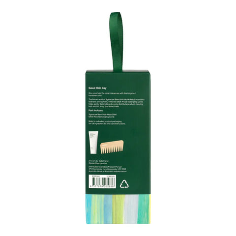 Back view of Good Hair gift packaging, showing a dark green box with a matching hanging loop, product text, a small tube icon, and a wooden comb illustration. The box has a striped multicolor base at the bottom and is photographed on a white background.