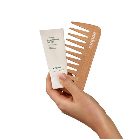 A hand holding a white tube of Endota Signature Blended Hair Mask next to a light natural wood hair comb with the Endota brand name printed on it, set against a solid white background.