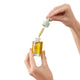 A hand holding a 30ml glass dropper bottle of endota Phyto Renewal Oil in one hand, while the other hand is holding the dropper cap. The bottle is prominently displayed with its label facing forward. The background is white.