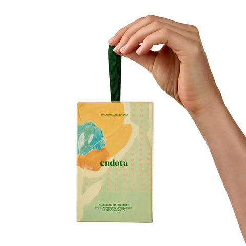 A hand holds a small rectangular product box with a dark green hanging loop against a plain white background. The box has abstract artwork in yellow, teal, and cream, with the visible brand name 'endota' printed near the bottom.