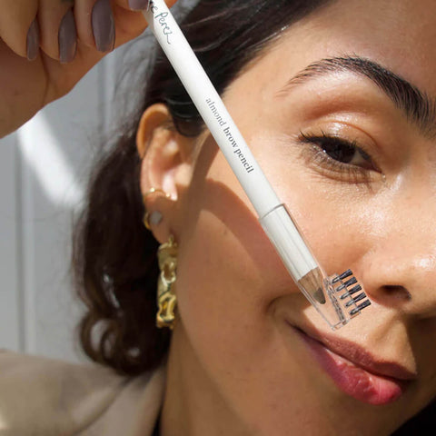 Ere Perez Almond Brow Pencil Perfect 1.1g lifestyle close up of a woman with a gold earring using the pencil and spoolie on her brow