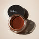 Open jar of Ere Perez Bronzing Pot -Costa 7.5g brown makeup product on a beige background.