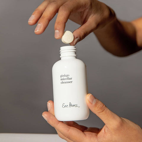 Person holding a white bottle labeled 'Ere Perez ginkgo micellar cleanser 37g' with a dropper, against a gray background.