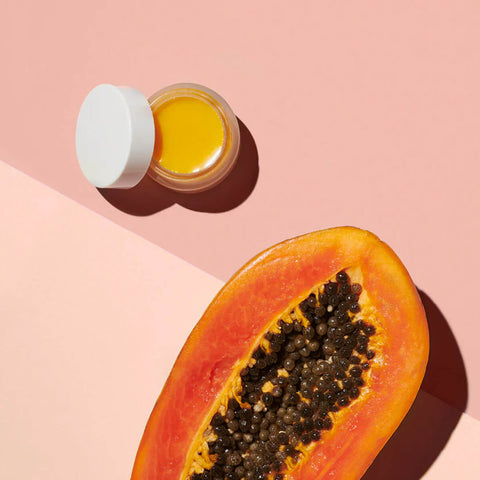Ere Perez Papaya SOS Marmalade 30g lifestyle shot of an open pot of balm beside half a papaya on a pink and peach backdrop, overhead