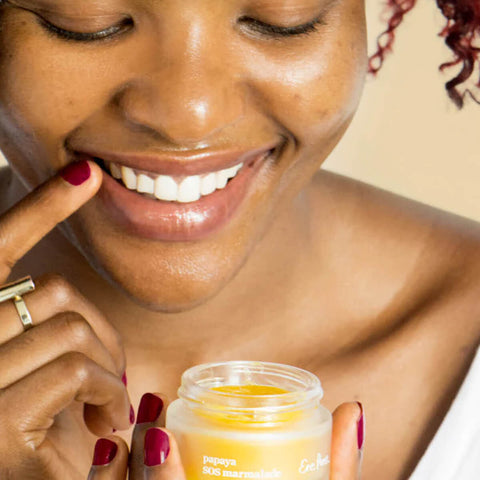 Ere Perez Papaya SOS Marmalade 30g lifestyle shot of a smiling woman with deep skin holding the open jar of balm near her chin