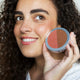 Ere Perez Rice Powder Blush & Bronzer Brooklyn lifestyle close up of a smiling woman with dark curly hair holding the open compact to her cheek