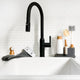 A matte black single-handle kitchen faucet with a high-arc pull-out spout installed in a white sink, set against a white subway tile backsplash. A soap dispenser, dish brush, and other bathroom accessories sit on a small tray to the right.