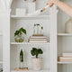 A hand uses a white duster with a wooden handle to clean a white bookshelf with potted plants and books.