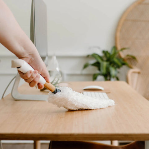 A hand holding a white microfiber duster with a wooden handle cleans a light wood desk in a bright, modern home office setting.