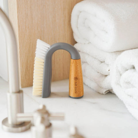 A small hand brush with a gray handle and light wood handle end, standing on a bathroom counter beside a faucet, with rolled white towels in the background.