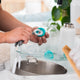 A close-up of a person's hands holding a small teal and white circular cleaning brush over a stainless steel sink, with bathroom items blurred in the background.