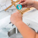 A close-up of a person's hands holding a small teal plastic cap and a white applicator wand, dispensing a white liquid onto a metal surface in a sink. A wooden stand is blurred in the background.