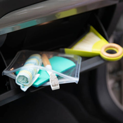 A clear zippered pouch containing a rolled towel, a toothbrush, and a turquoise sponge, resting in a car glove box next to a lime green hand mirror.