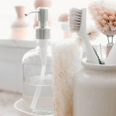 A close-up of bathroom cleaning and soap accessories, including a clear glass pump bottle with a silver dispenser, a white-handled brush, a beige scrubber, and a white container holding cleaning tools, set in a bright, softly blurred bathroom scene.