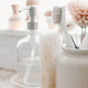 A close-up of bathroom cleaning and soap accessories, including a clear glass pump bottle with a silver dispenser, a white-handled brush, a beige scrubber, and a white container holding cleaning tools, set in a bright, softly blurred bathroom scene.