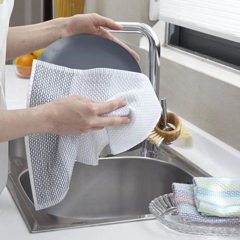 A person's hands holding a dark gray plate while drying it with a white textured dish towel with black woven dots in a modern kitchen sink setting.
