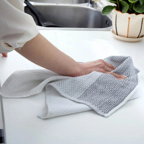 A person's hand and arm are shown cleaning a white kitchen countertop with a gray and white textured microfiber cloth. In the background, a stainless steel sink and a potted green plant are visible.